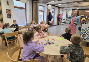 Dzieci siedzą przy okrągłych stolikach i słuchają Pani bibliotekarki, która tłumaczy im sposób wykonania papierowej sowy.