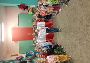 Dzieci stoją na baczność w sali zabaw i śpiewają Hymn Polski. Kilkoro z nich trzyma dużą flagę Polski.