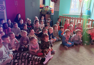 Dzieci siedzą na dywanie i oglądają przedstawienie teatralne.