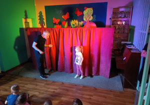 Dzieci oglądają przedstawienie teatralne. Dziewczynka stoi na scenie i słucha instrukcji aktora.
