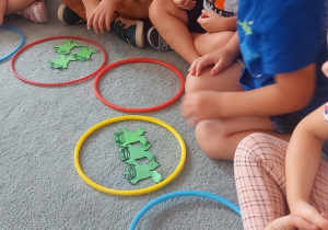 Dzieci siedzą na dywanie maja przed sobą obręcze a w nich papierowe żabki.