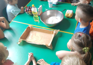 Dzieci siedzą dookoła stołu, na którym ustawione są wszystkie niezbędne produkty. Dzieci wymieniają składniki potrzebne do upieczenia ciasta.