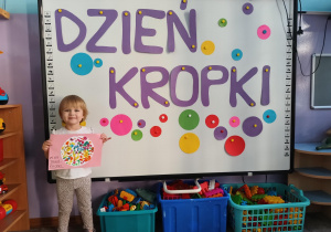 Dziewczynka pozuje do zdjęcia na tle ozdobionej tablici i prezentuje swoją pracę plastyczną.