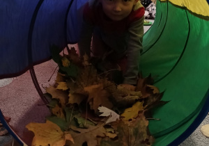 Dziewczynka przechodzi przez tunel wysypany jesiennymi liśćmi.