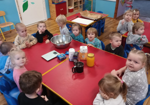 Dzieci siedzą przy stoliku, na którym leżą przygotowane produkty potrzebne do upieczenia ciasteczek dyniowych.