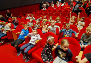 Zdjęcie dzieci siedzących przed przedstawieniem na fotelach, na widowni.