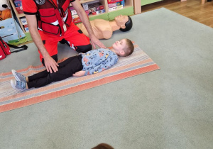 Chłopiec leży przed ratownikiem, który pokazywać będzie jak ułożyć poszkodowanego w pozycji bezpiecznej.