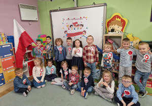 Dzieci pozują do zdjęcia na tle tablicy z napisem Dzień Niepodległości. Do bluzek przyczepione mają własnoręcznie wykonane kotyliony. Jedno dziecko trzyma flagę Polski.
