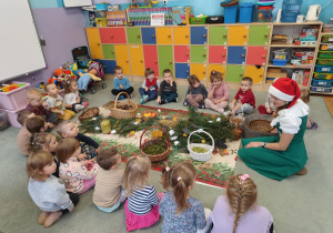 Dzieci siedzą na dywanie dookoła świątecznego obrusu. A na nim: gałązki choinkowe, koszyki z orzechami, szyszkami, laskami cynamonu, rozłożone kwiaty lawendy. plasty pomarańczy