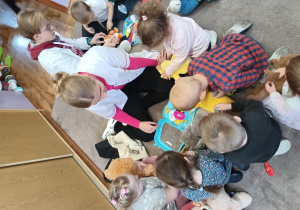 Dzieci w małych grupach wraz ze studentami siedzą na dywanie i poznają wyposażenie zabawkowego zestawu lekarskiego.