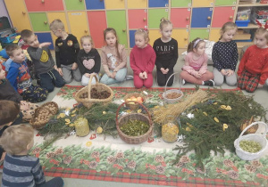 Dzieci siedzą na dywanie dookoła świątecznego obrusu. A na nim: gałązki choinkowe, koszyki z orzechami, szyszkami, laskami cynamonu, rozłożone kwiaty lawendy, plasty pomarańczy.