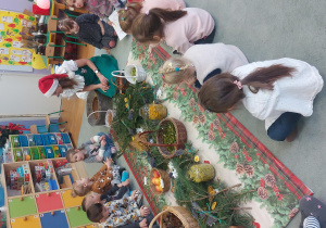 Dzieci siedzą na dywanie dookoła świątecznego obrusu. A na nim: gałązki choinkowe, koszyki z orzechami, szyszkami, laskami cynamonu, rozłożone kwiaty lawendy, plastry pomarańczy.