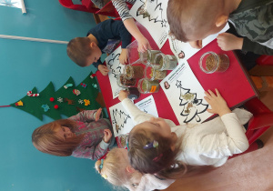 Dzieci siedzą przy stoliku i wykonują ziołowa choinkę. Posypują choinkę różnymi ziołami.