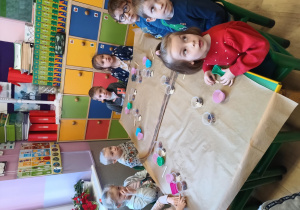 Dzieci siedzą dookoła stołu, na którym przygotowane mają foremki na masę mydlaną oraz naturalne składniki, które dodadzą do mydła
