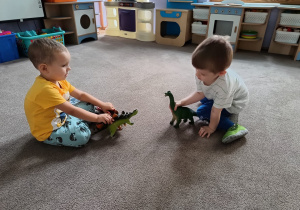 dwoje chłopców bawi się na dywanie dinozaurami.