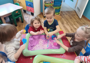 Dzieci siedzą przy stoliku i wspólnie bawią się piaskiem kinetycznym.