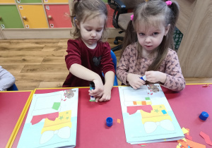 Dwie dziewczynki przyklejają małe kawałki kolorowego papieru na rysunek prezentu.