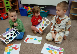 Dzieci siedzą na dywanie i wypychają z kartony różne kształty. Przed nimi leży kartka na której je układają.