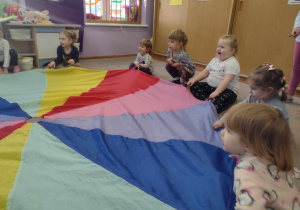 Dzieci siedzą na dywanie i trzymając chustę aminacyjną słuchają objaśnień nauczycielki