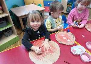 Dziewczynka siedzi przy stoliku i maluje czerwoną kredką spód papierowej pizzy.
