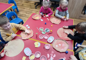 Dzieci siedzą przy stoliku i naklejają na pomalowaną pizzę różne produkty. Każde wg własnego uznania.