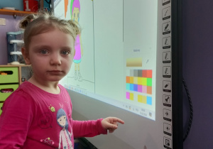 Dziewczynka stoi przed tablicą multimedialną i wybiera kolor, na który zamaluje część obrazka.