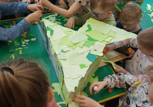 Dzieci obklejają szkielet dinozaura kawałkami zielonego papieru