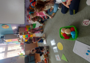 Dzieci siedzą na dywanie w kole i rozwiązują zadania matematyczne.