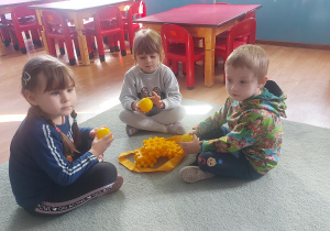 Troje dzieci siedzi na dywanie. W rękach trzymają żółte piłki.