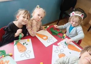 Dzieci siedzą przy stoliku i wklejają kolorowym papierem sylwetę marchewk.