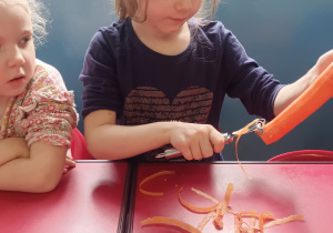 Dziewczynka siedzi przy stoliku i obiera marchew przy pomocy obieraczki.