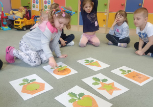 Dzieci siedzą na dywanie. Dziewczynka dopasowuje odpowiedni kształt do kształtu marchewki.