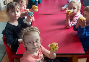 Dzieci siedzą przy stolikach i pokazują babeczki, które sami przygotowali. Zaraz będą mogli je zjeść.