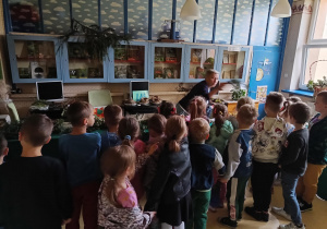 Dzieci stoja w bibliotece szkolnej tyłem do obiektywu i słuchają Pani bibliotekarki która, opowiada o darach jesieni.