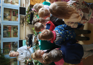 Dzieci stoją tyłem do obiektywu w bibliotece szkolnej i słuchają Pani bibliotekarki, która opowiada o darach jesieni.