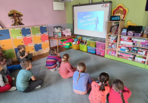 Dzieci siędzą na dywanie i oglądają film edukacyjny wyświetlany na tablicy multimedialnej.