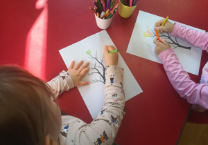 Dzieci przy stolikach dorysowują wydrukowanym pniom drzew, jesienne liście.