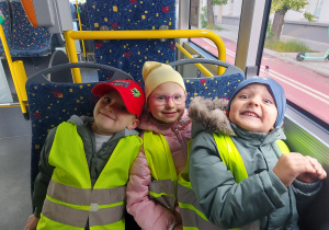 Troje dzieci w kamizelkach odblaskowych siedzi na krzesłach w autobusie.