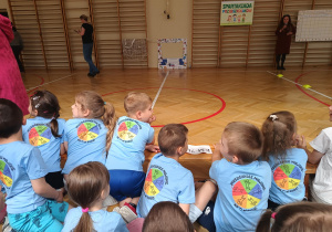 Przedszkolaki w koszulkach z logo przedszkola siedzą na podłodze w szkolnej sali gimnastycznej.