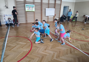 Grupa przedszkolaków bierze udział w konkurencji sportowej na sali gimnastycznej.