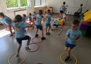 Przedszkolaki w koszulkach z logo przedszkola wykonują zadania sportowe.
