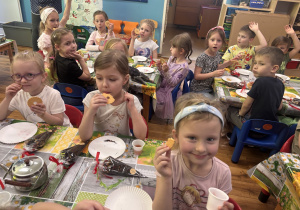 Dzieci siedzą przy stolikach, jedzą ciastka i popijają herbatą.