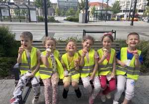 Trzech chłopców i trzy dziewczynki w kamizelkach odblaskowych jedzą lody siedząc na ławce.