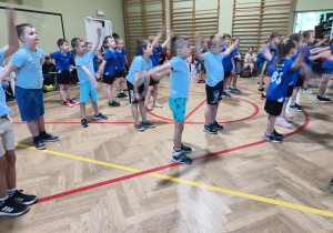 Grupa przedszkolaków w niebieskich podkoszulkach wykonują ćwiczenia na sali gimnastycznej.