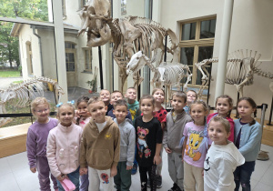 Grupa dzieci pozuje do zdjęcia na tle szkieletów dinozaurów.
