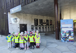Grupa dzieci ubrana w kamizelki odblaskowe stoi przed wejściem do Zoo-Orientarium.
