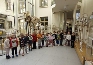 Dzieci pozują do zdjęcia w sali ze szkieletami dinozaurów.