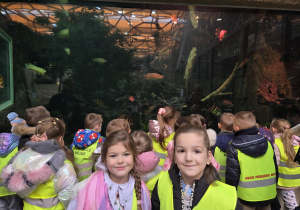 Dwie dziewczynki pozują do zdjęcia. Za nimi tyłem stoi grupa dzieci, która ogląda ryby pływające w akwarium.