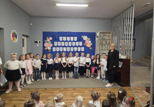 Dzieci ubrane na galowo stoją w sali przedszkolnej. Za nimi na tablicy napis: Dzień Edukacji Narodowej.