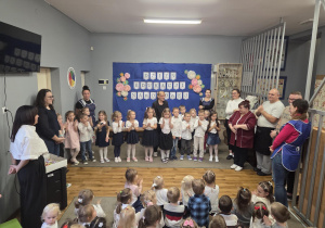 Dzieci z grupy 3 stoją w sali przedszkolnej razem z pracownikami przedszkola.